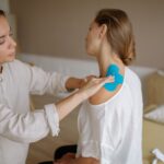 A physiotherapist applies blue kinesiology tape to a patient's shoulder for muscle support.